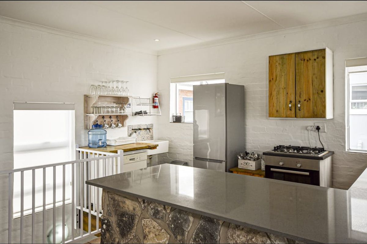 A compact kitchen featuring a stone-clad breakfast bar, built-in oven and gas hob, white painted brick walls, and a farmhouse-style sink.