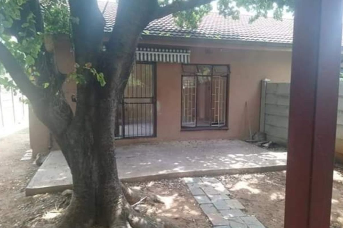 Rear exterior view of the property featuring a concrete patio, security gate, and a large tree in the garden area.
