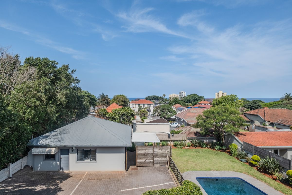 Aerial view of the property featuring a paved driveway, swimming pool, and landscaped garden with surrounding residential neighborhood and distant sea view.