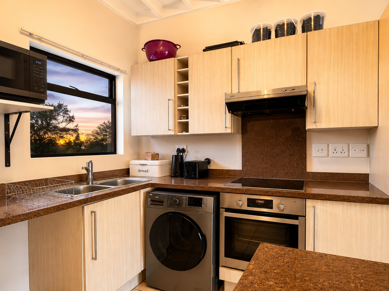 Modern kitchen featuring light wood cabinetry, stone countertops, built-in oven, hob, extractor fan, and integrated washing machine with a scenic window view.