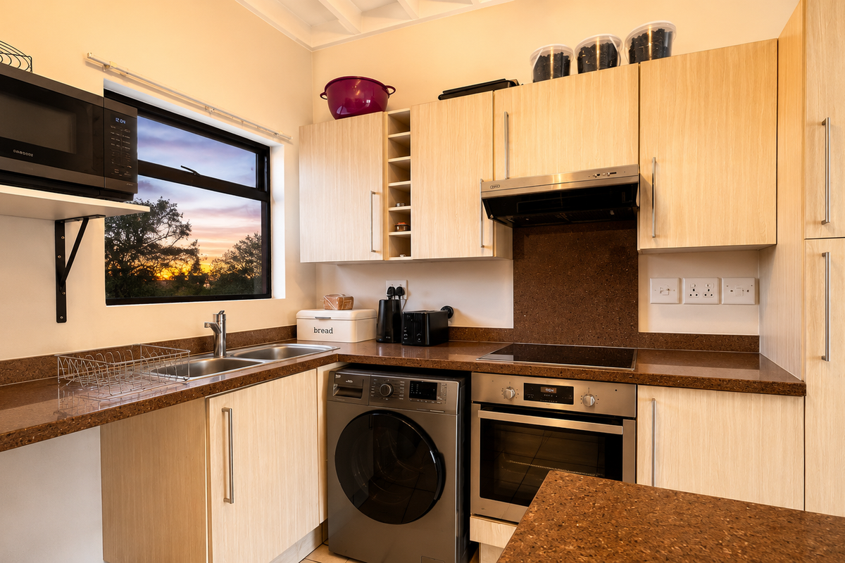 Modern kitchen featuring light wood cabinetry, stone countertops, built-in oven, hob, extractor fan, and integrated washing machine with a scenic window view.