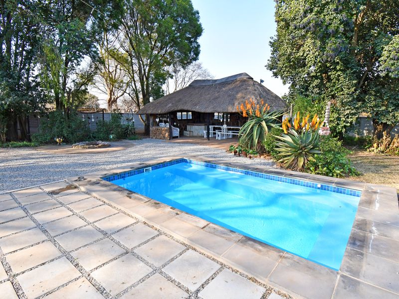 Outdoor entertainment area featuring a swimming pool with paved surround and a traditional thatched lapa set within a landscaped garden.