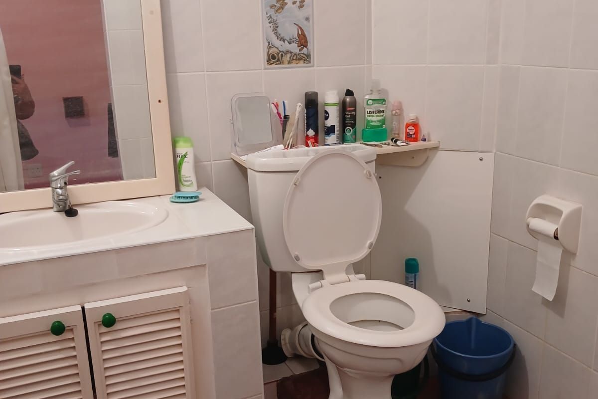 A functional bathroom featuring a vanity cabinet with basin, wall-mounted mirror, and standard toilet, finished with white wall tiles and terracotta floor tiles.
