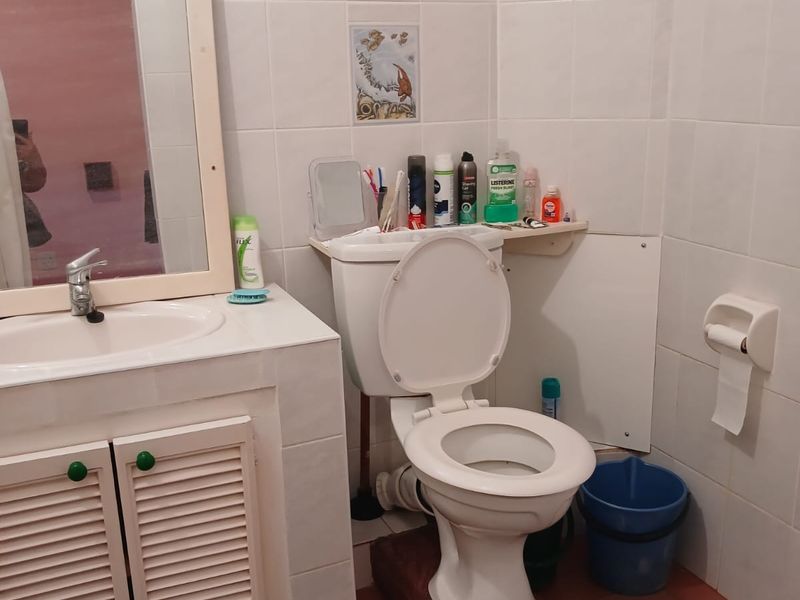 A functional bathroom featuring a vanity cabinet with basin, wall-mounted mirror, and standard toilet, finished with white wall tiles and terracotta floor tiles.