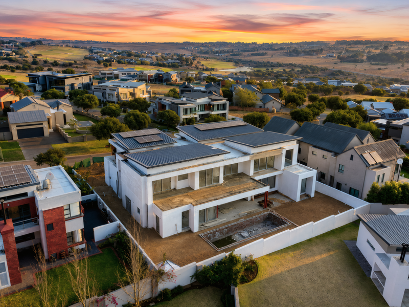 Aerial view of a modern residential property under construction, featuring solar panels, a swimming pool, and surrounding estate landscape at sunset.