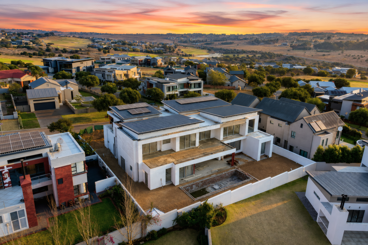Aerial view of a modern residential property under construction, featuring solar panels, a swimming pool, and surrounding estate landscape at sunset.