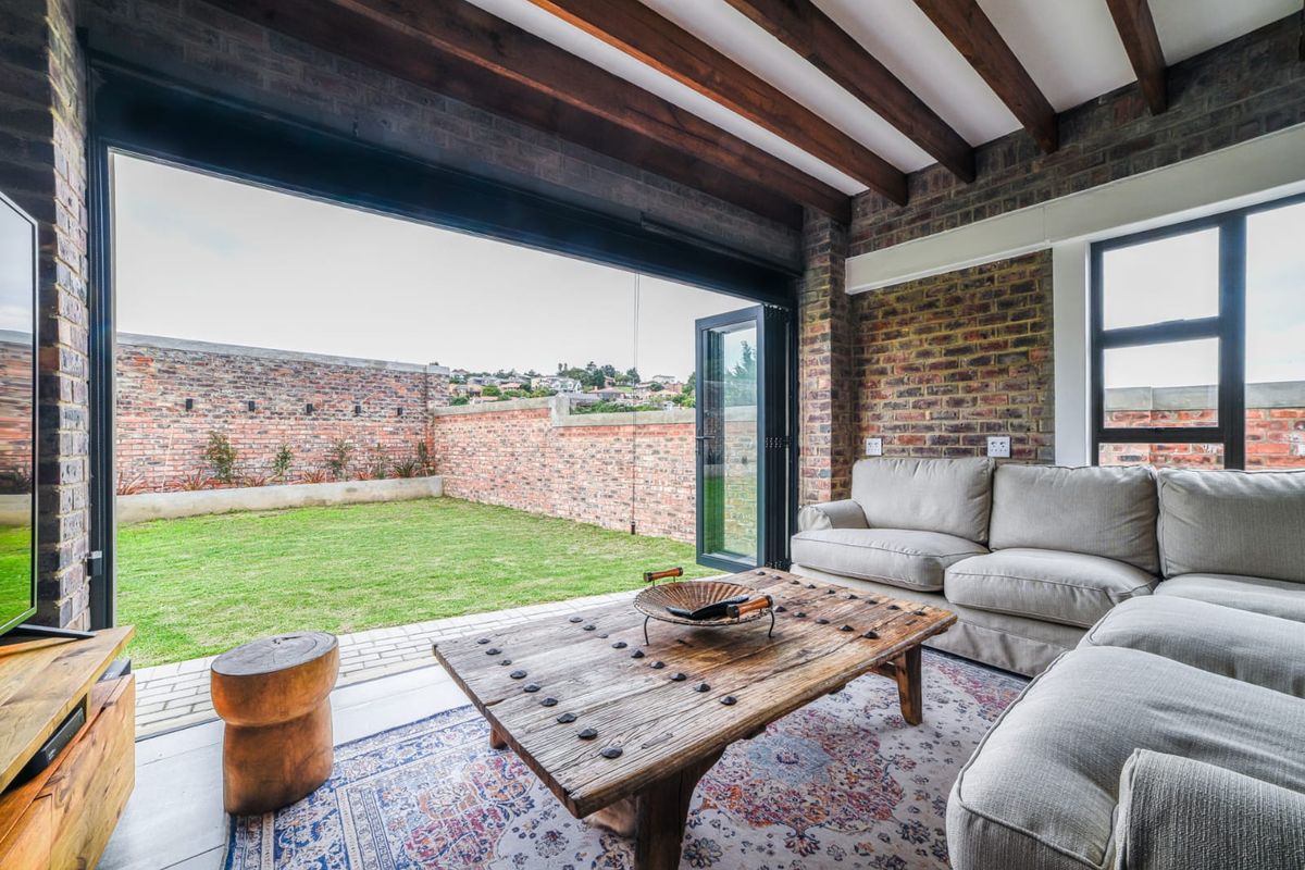 A bright living room featuring exposed brick walls, timber ceiling beams, and large folding doors that open onto a private garden.