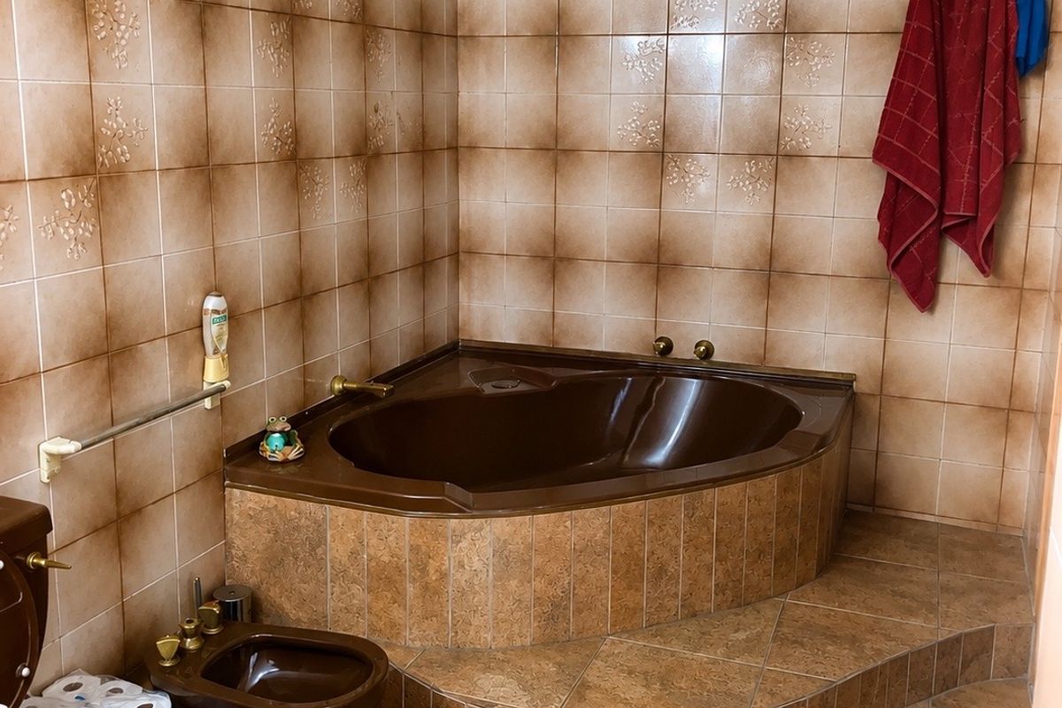 A dated bathroom featuring a built-in corner bathtub, bidet, and toilet with matching brown sanitaryware and extensive ceramic wall and floor tiling.