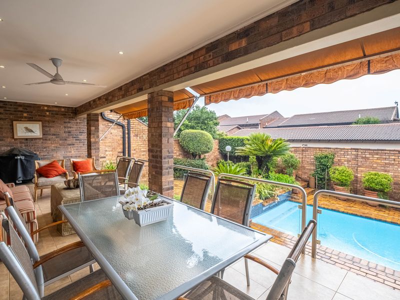 Covered outdoor patio featuring exposed brickwork, tiled flooring, and a glass balustrade overlooking a private swimming pool and landscaped garden area.