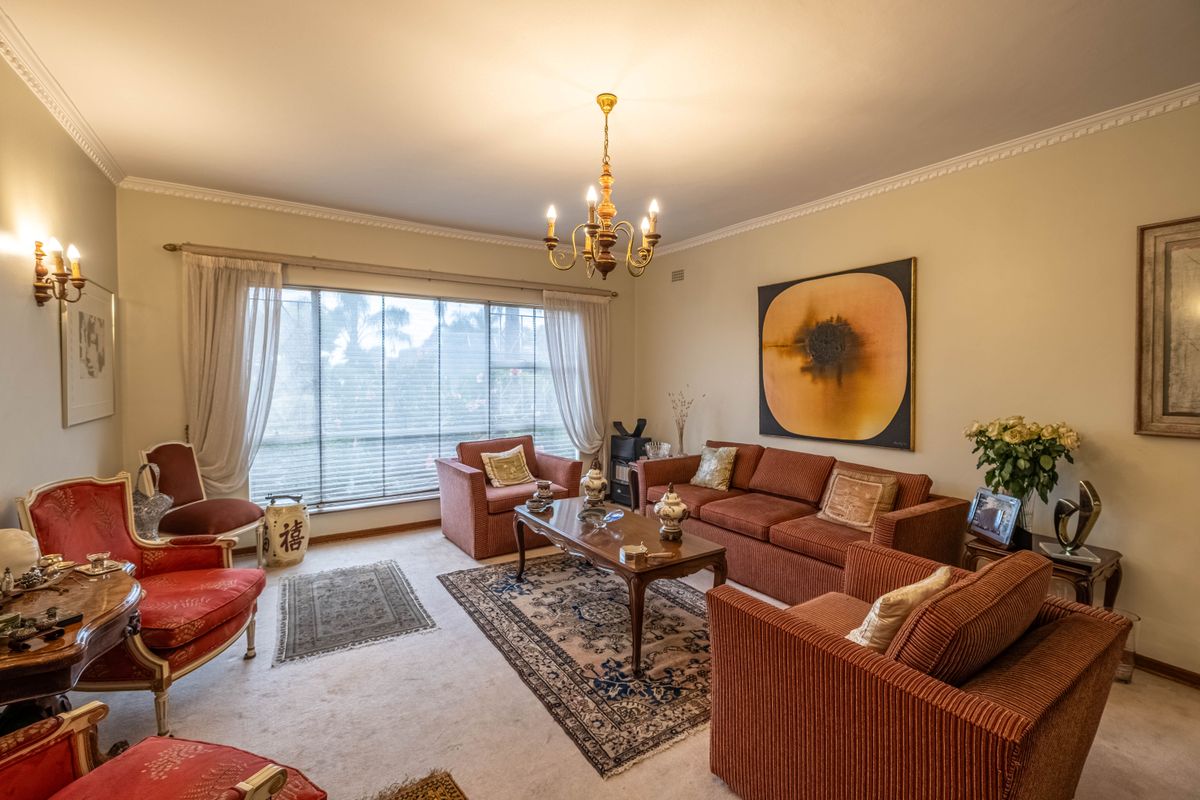 A spacious living room featuring crown molding, a classic chandelier, wall sconces, and a large window allowing natural light into the carpeted space.