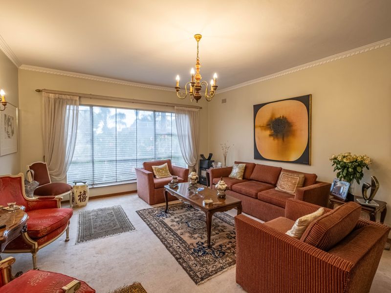 A spacious living room featuring crown molding, a classic chandelier, wall sconces, and a large window allowing natural light into the carpeted space.