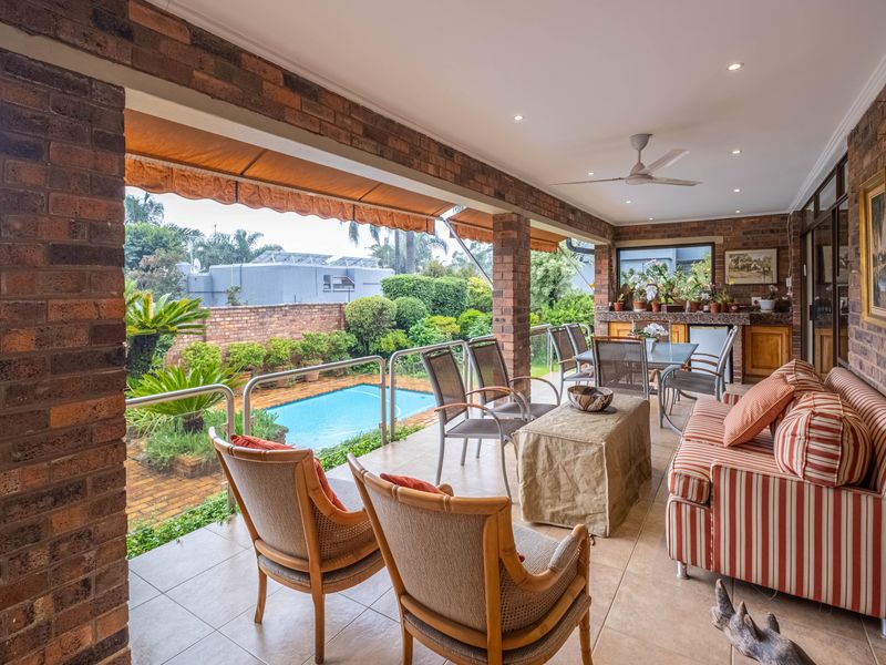 A spacious covered patio featuring exposed brick pillars, tiled flooring, a ceiling fan, and a built-in outdoor counter, overlooking the garden and pool area.