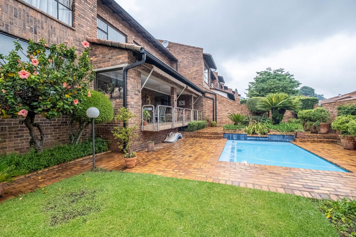 Rear exterior view of a brick townhouse featuring a paved patio, covered veranda with glass balustrade, and a private swimming pool set in a garden.