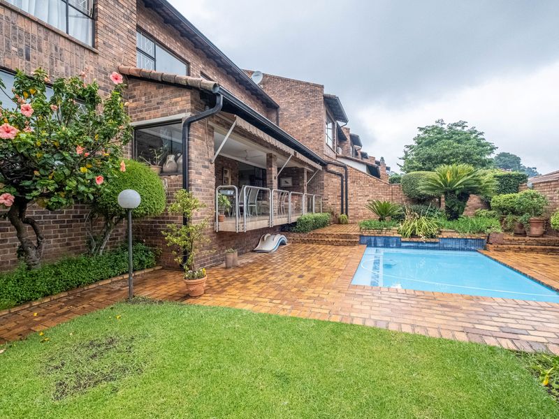 Rear exterior view of a brick townhouse featuring a paved patio, covered veranda with glass balustrade, and a private swimming pool set in a garden.