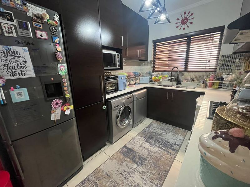 A functional kitchen featuring dark wood cabinetry, integrated appliances including a washing machine and dishwasher, double sink, and tiled backsplash with venetian blinds.