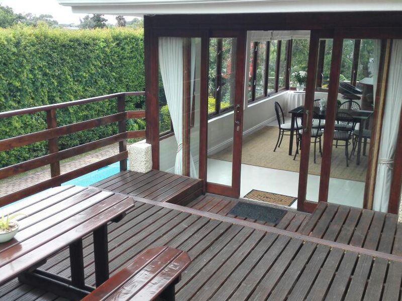 Spacious timber deck featuring a wooden balustrade, overlooking a pool area and providing access to an enclosed sunroom through sliding glass doors.