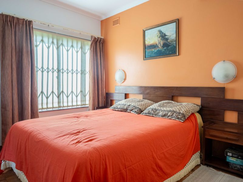 A bedroom featuring parquet flooring, built-in headboard, and wall-mounted lighting, with a window protected by a security gate.