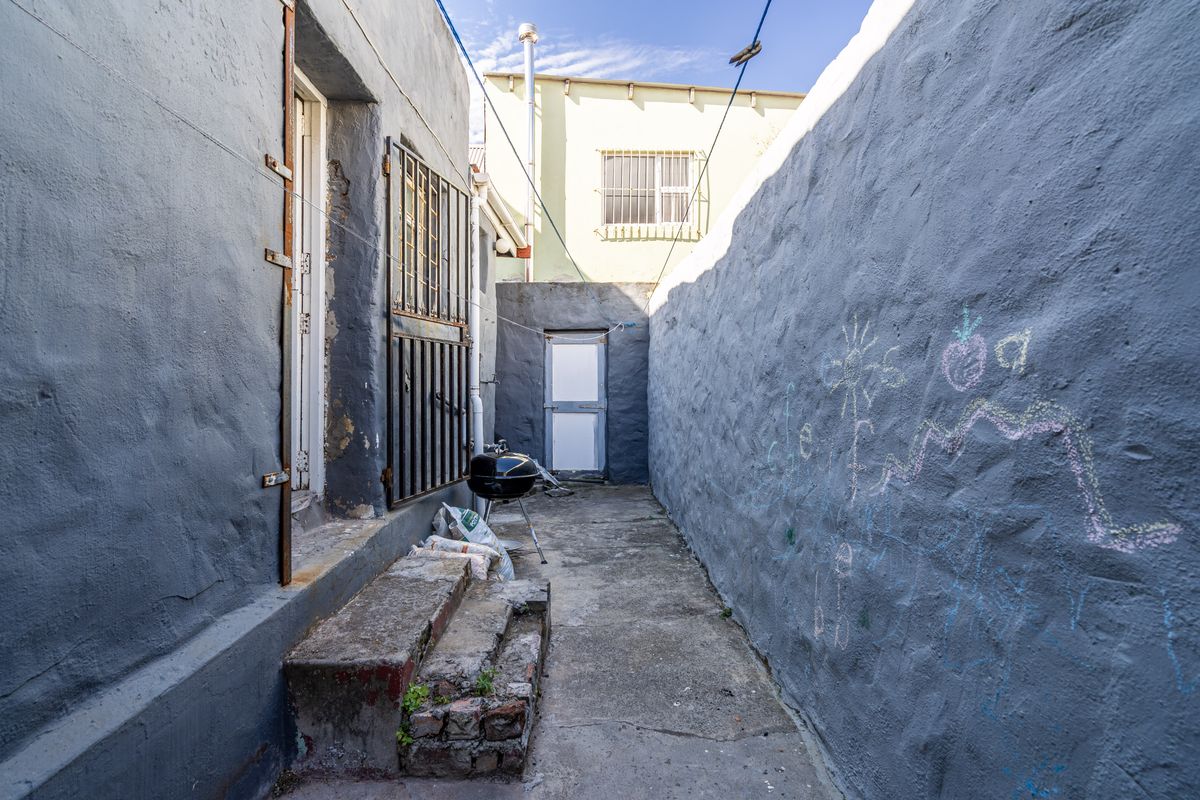 A narrow rear exterior passage featuring textured masonry walls, concrete steps, and a security gate, requiring maintenance and cosmetic updates.