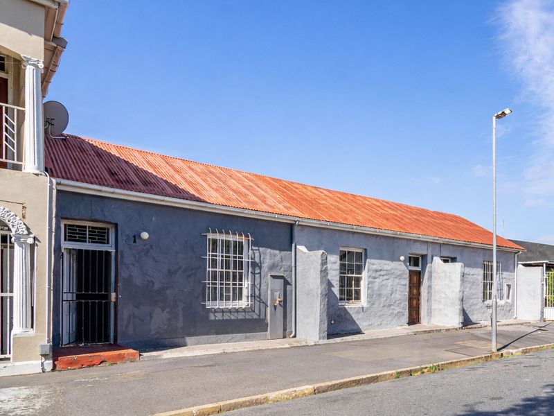 Street-facing view of a single-story residential property featuring a corrugated metal roof, security-barred windows, and plastered exterior walls.