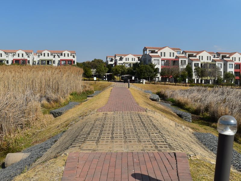 A paved walkway leading through a landscaped estate area with gabion retaining walls and views of the surrounding apartment complex buildings.