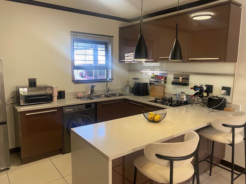 Modern kitchen featuring high-gloss brown cabinetry, stone countertops, a gas hob, integrated washing machine, and a breakfast bar with pendant lighting.