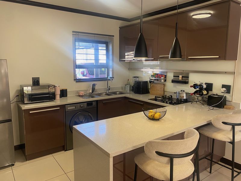 Modern kitchen featuring high-gloss brown cabinetry, stone countertops, a gas hob, integrated washing machine, and a breakfast bar with pendant lighting.