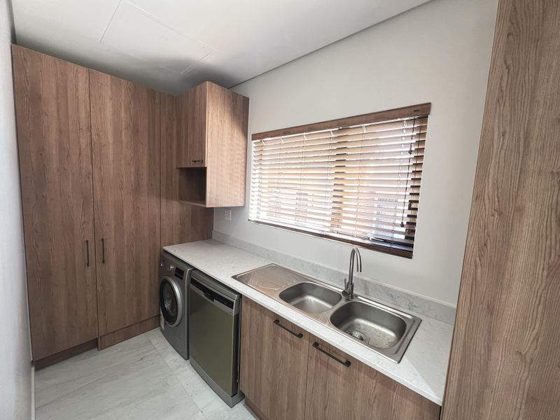 A modern, well-maintained scullery featuring wood-finish cabinetry, stone countertops, a double stainless steel sink, and integrated laundry and dishwashing appliances.