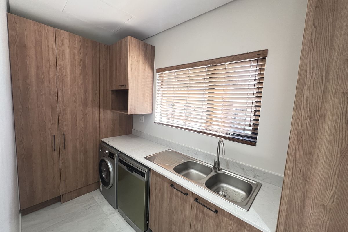 A modern, well-maintained scullery featuring wood-finish cabinetry, stone countertops, a double stainless steel sink, and integrated laundry and dishwashing appliances.