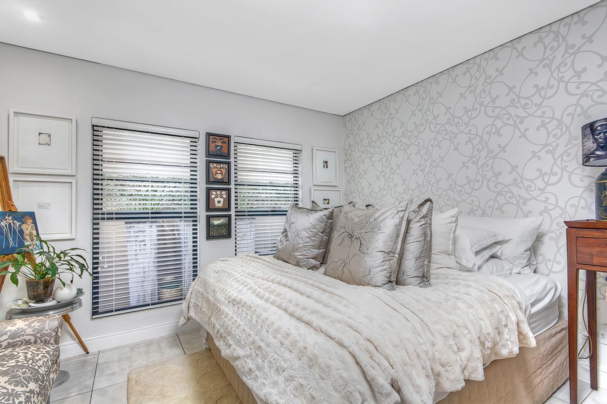 A well-maintained bedroom featuring neutral tones, patterned accent wallpaper, tiled flooring, and dual windows fitted with venetian blinds.