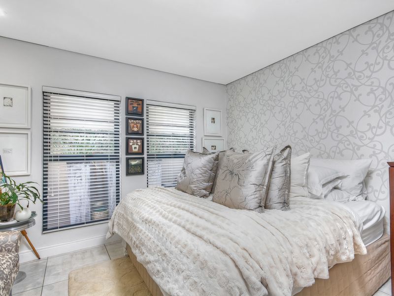 A well-maintained bedroom featuring neutral tones, patterned accent wallpaper, tiled flooring, and dual windows fitted with venetian blinds.