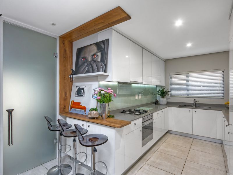 Modern kitchen featuring sleek white cabinetry, stone countertops, integrated gas hob, built-in oven, and a breakfast bar with wood accent shelving.