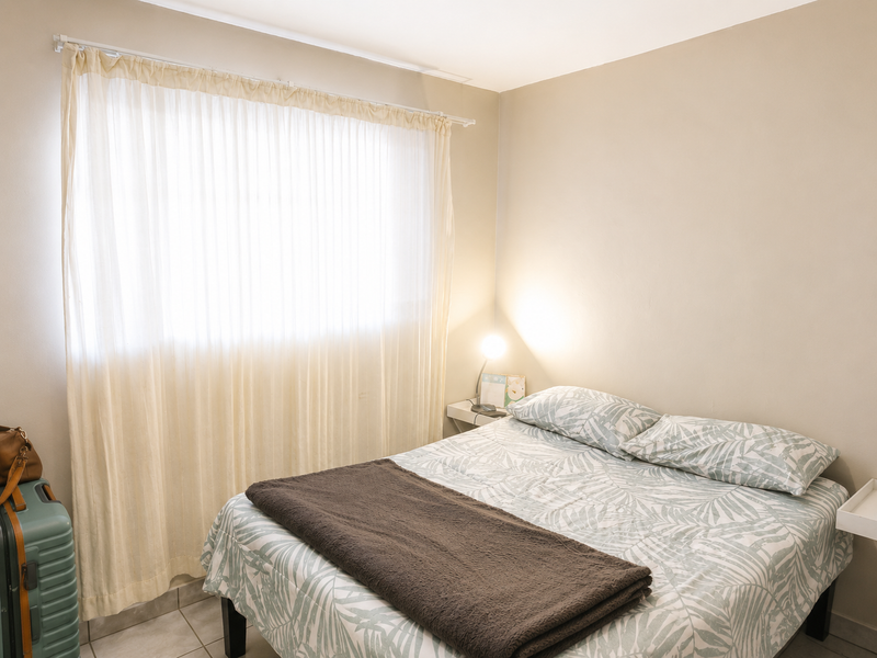 A functional bedroom featuring tiled flooring and a large window with a curtain rod, providing a neutral base for interior design updates.