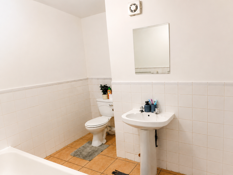 A functional bathroom featuring a pedestal sink, toilet, wall-mounted mirror, and extractor fan, finished with neutral ceramic wall and floor tiles.