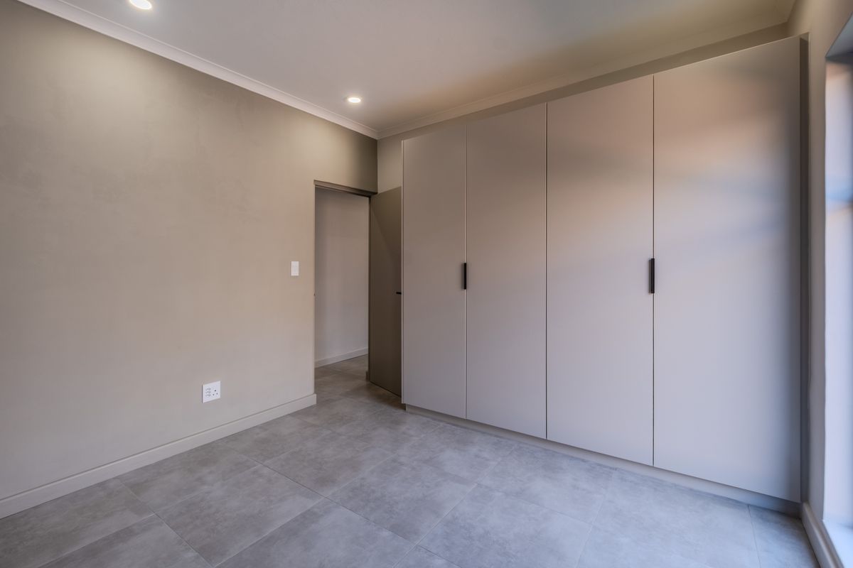 A modern, empty bedroom featuring neutral-toned walls, large built-in cupboards, recessed downlights, and contemporary grey floor tiles.