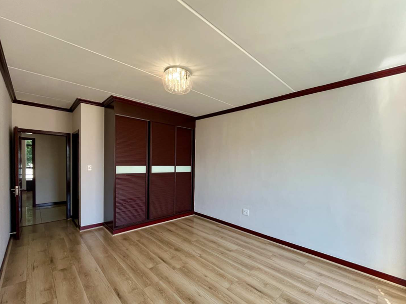 A bright, empty bedroom featuring modern laminate flooring, dark wood-finish built-in cupboards, and a central ceiling light fixture.