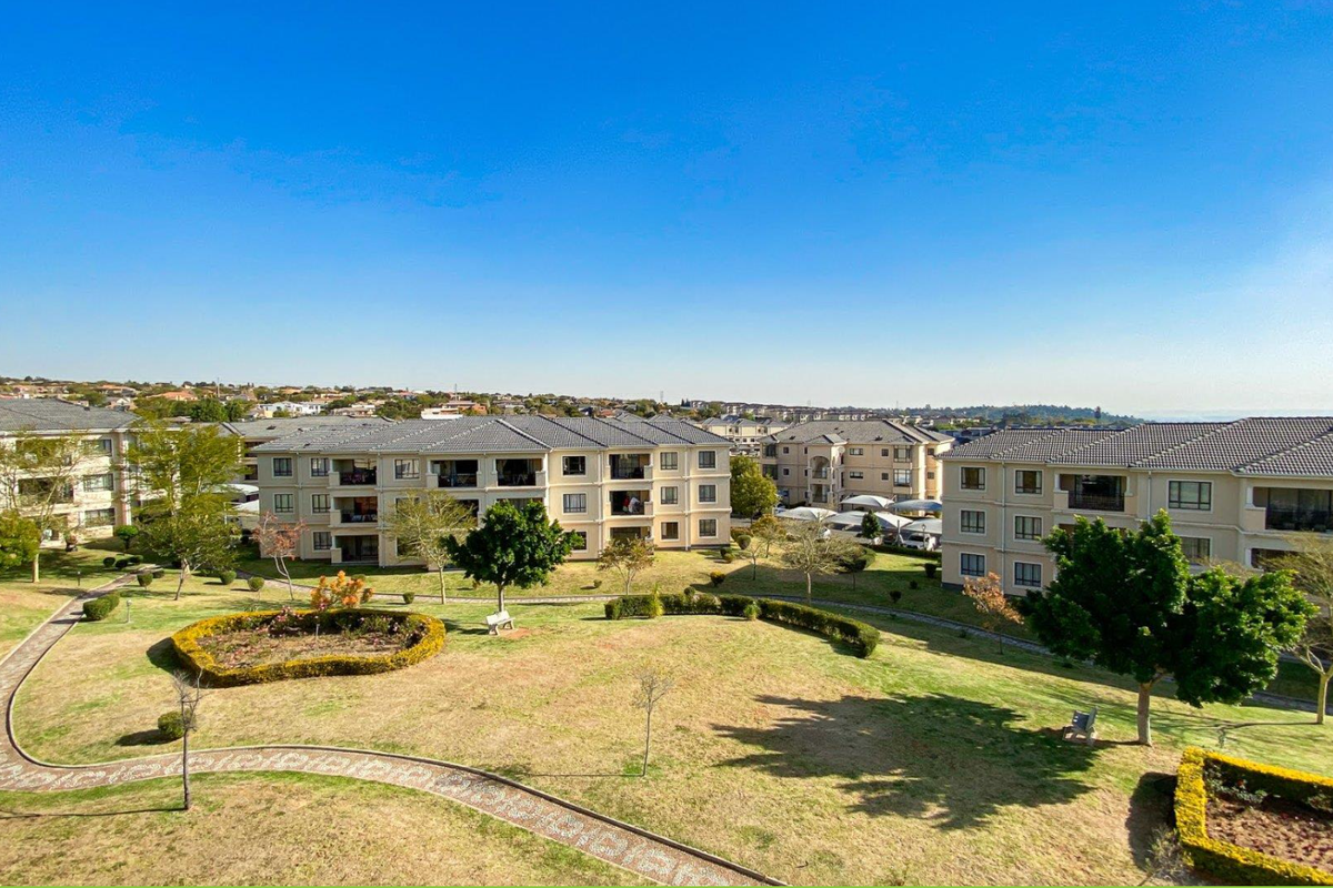 An elevated aerial view of a residential apartment complex featuring well-maintained landscaped gardens, paved pathways, and multiple apartment buildings with tiled roofs.