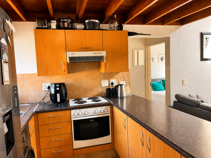 A functional kitchen featuring wooden cabinetry, laminate countertops, built-in oven and hob, and exposed timber ceiling beams, with a view into the adjacent bedroom.