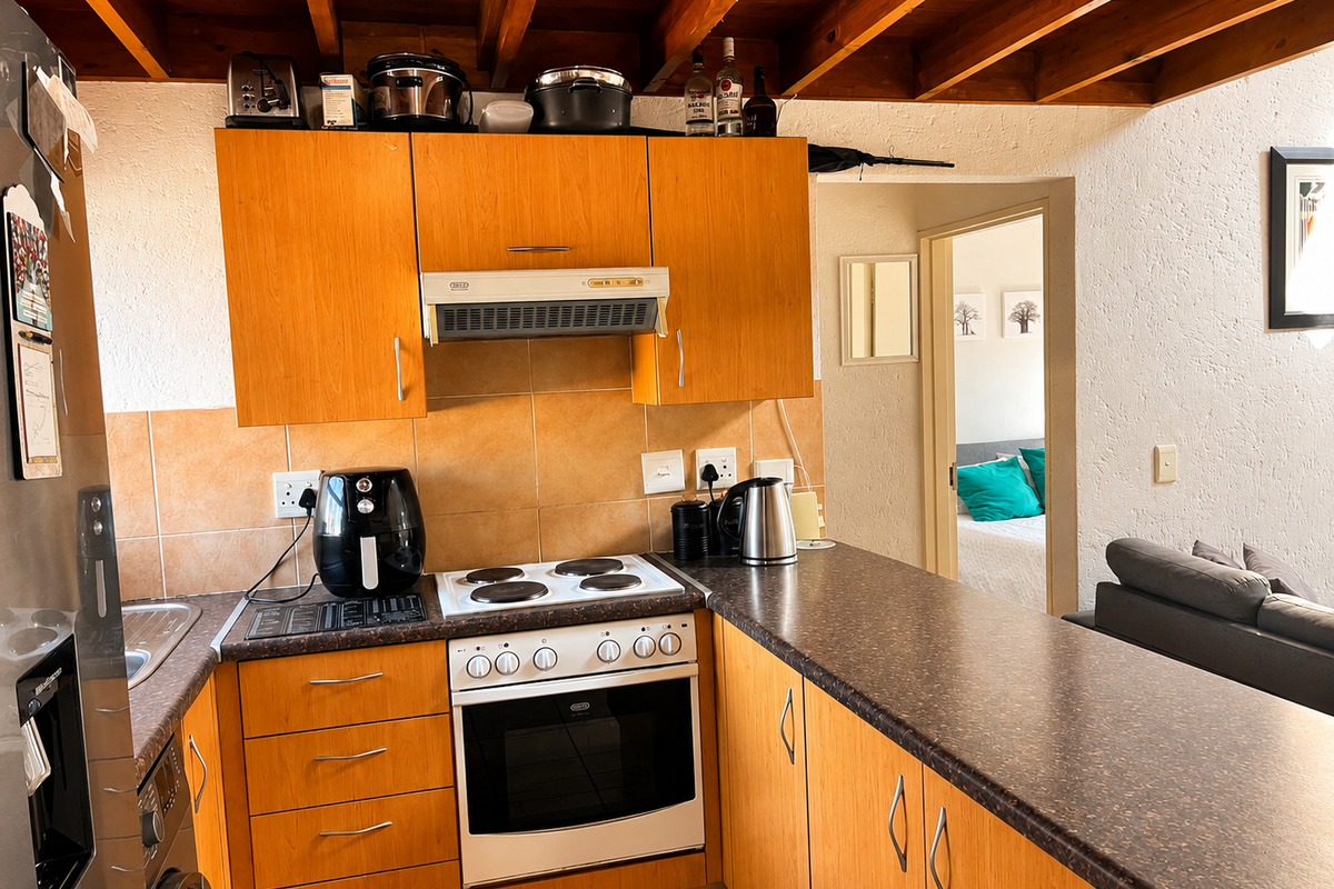 A functional kitchen featuring wooden cabinetry, laminate countertops, built-in oven and hob, and exposed timber ceiling beams, with a view into the adjacent bedroom.