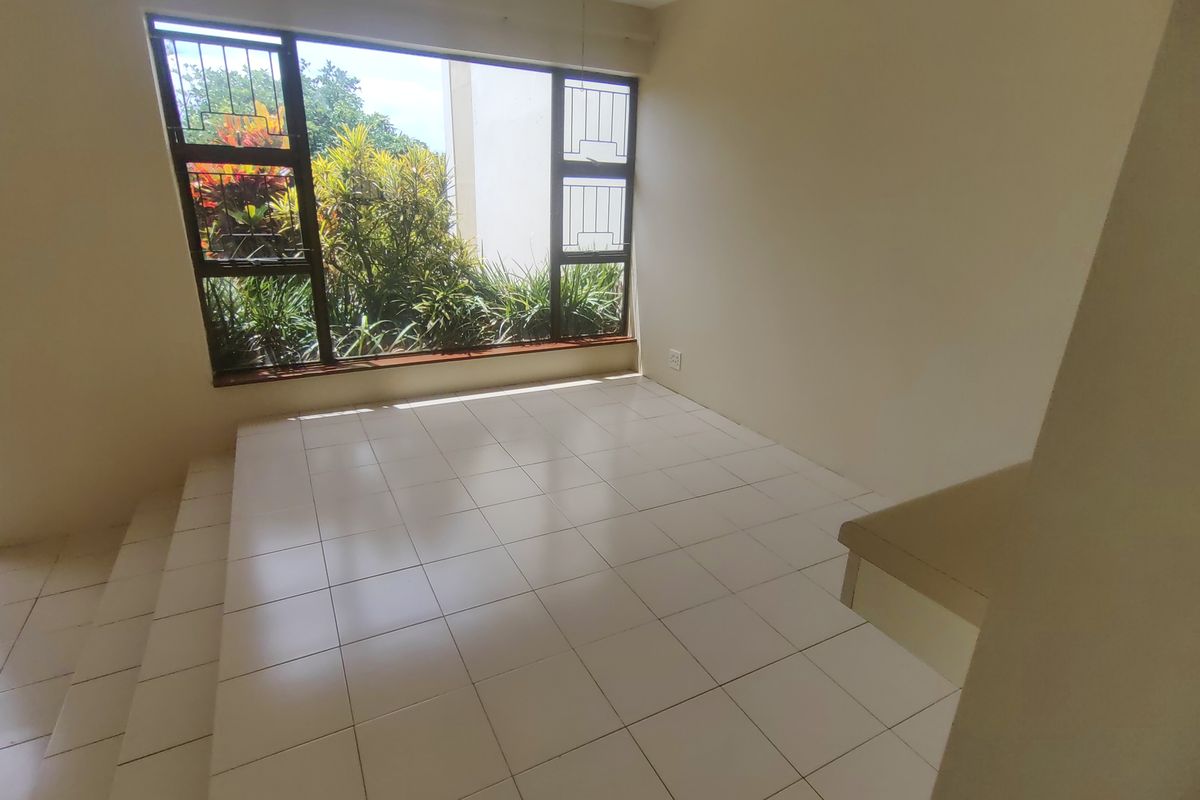 An empty living area featuring tiled flooring, a raised platform, and a large window with security bars overlooking garden greenery.
