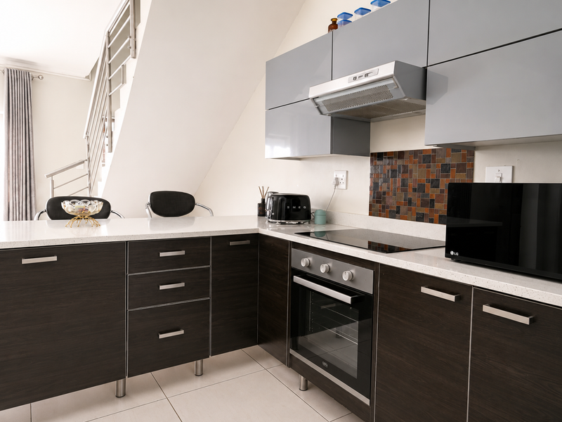 Modern kitchen featuring dark wood cabinetry, stone countertops, built-in oven, electric hob, and a tiled backsplash, situated adjacent to an internal staircase.