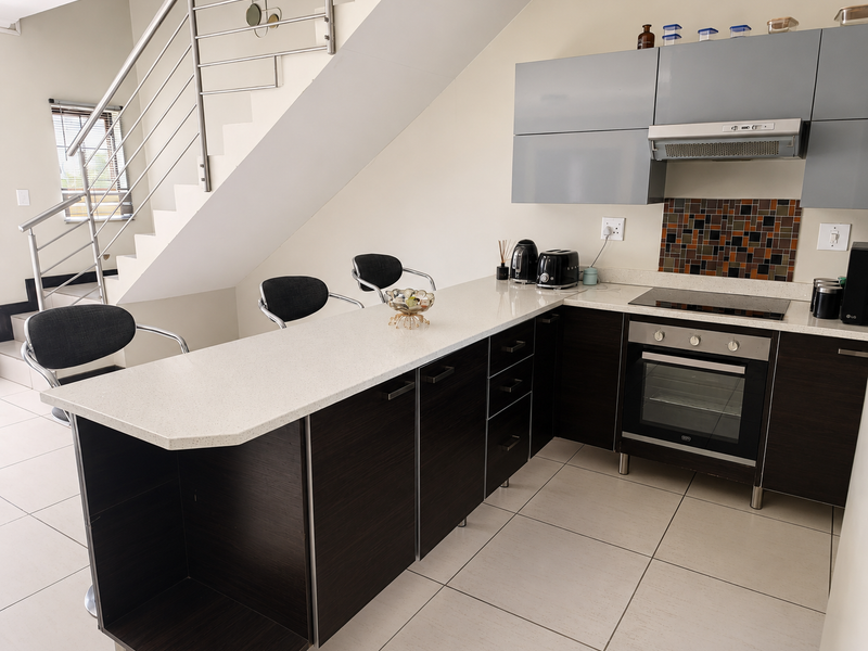Modern kitchen featuring dark cabinetry, quartz countertops, built-in oven, and hob, situated beneath a staircase with a contemporary stainless steel railing.