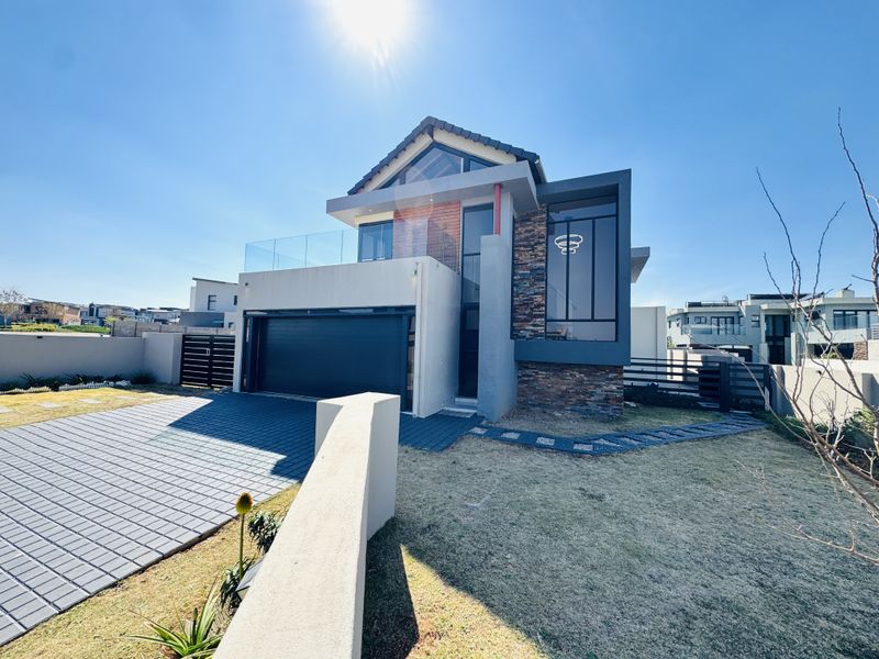Modern double-story residential home featuring stone cladding, a paved driveway, double garage, and a glass-railed balcony under a clear blue sky.