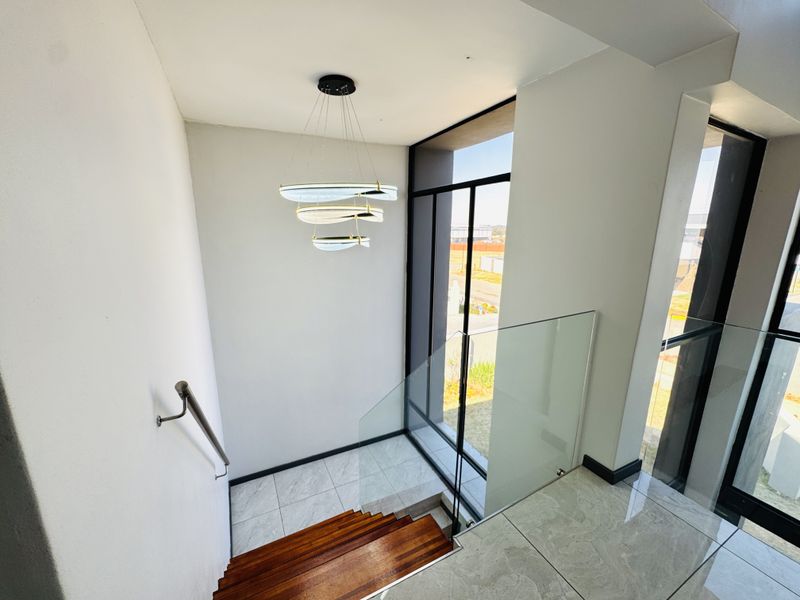 A bright, modern staircase area featuring timber treads, a sleek glass balustrade, and a contemporary pendant light fixture with large windows.