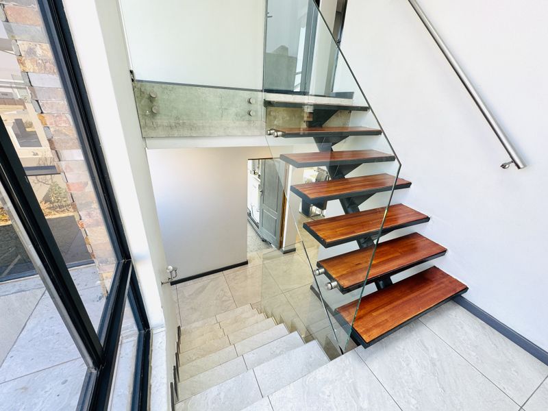 Modern architectural staircase featuring timber treads, glass balustrades, and stainless steel handrails, set against a bright, tiled hallway with large windows.