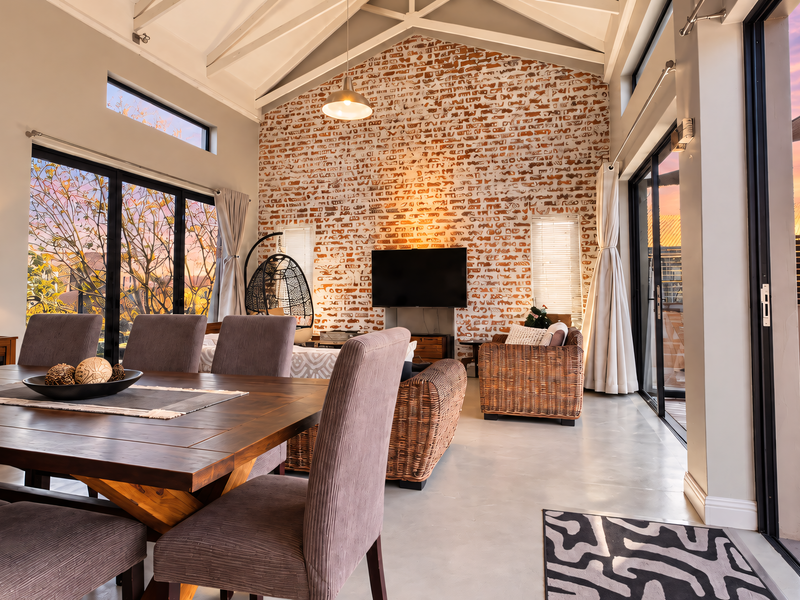 Spacious open-plan living and dining area featuring a striking exposed brick wall, vaulted ceilings with exposed rafters, and seamless access to an outdoor deck.