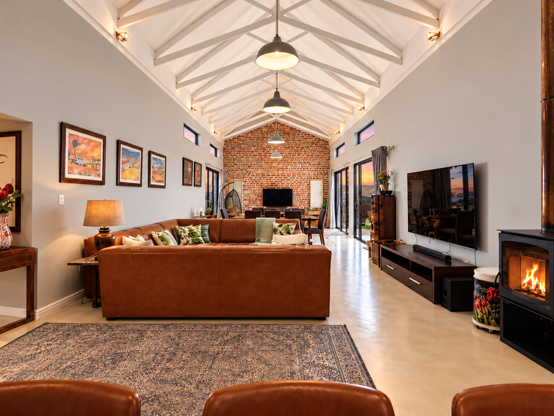 Spacious open-plan living area featuring a vaulted ceiling with exposed timber trusses, polished concrete floors, and a striking exposed brick feature wall with fireplace.
