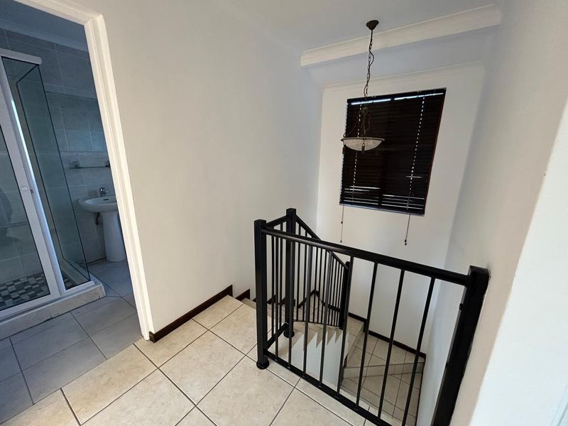 A bright hallway landing featuring tiled flooring, a metal staircase balustrade, and direct access to an adjacent bathroom with a shower and pedestal basin.