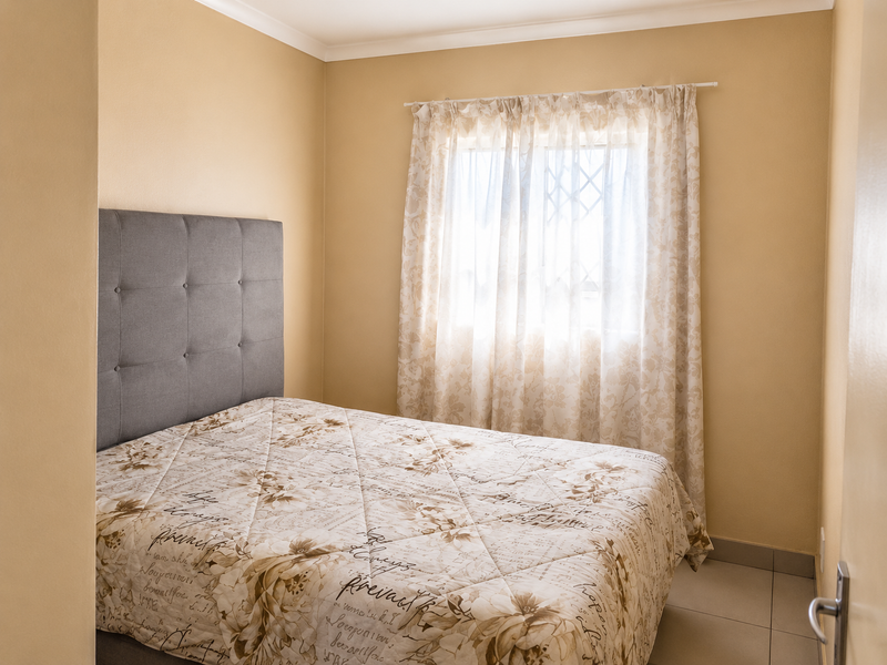 A compact bedroom featuring neutral wall tones, tiled flooring, and a window fitted with security bars.