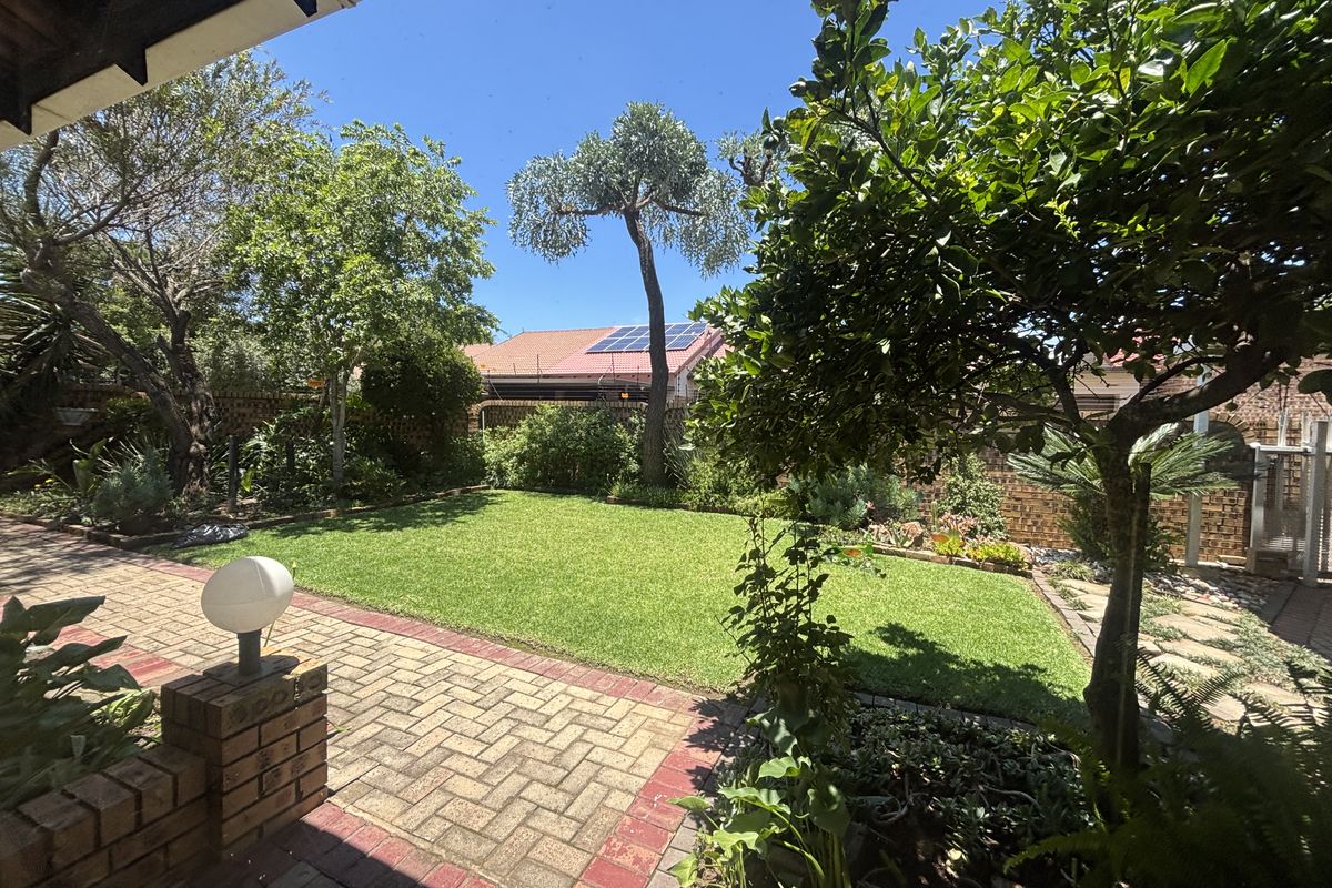 A well-maintained rear garden featuring a paved patio area, lush green lawn, mature trees, and perimeter brick walling.