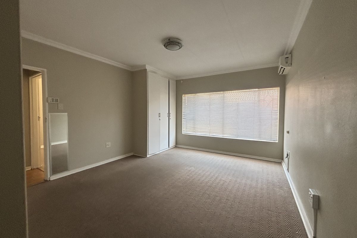 A spacious, empty bedroom featuring neutral wall tones, built-in cupboards, carpeted flooring, and a wall-mounted air conditioning unit.
